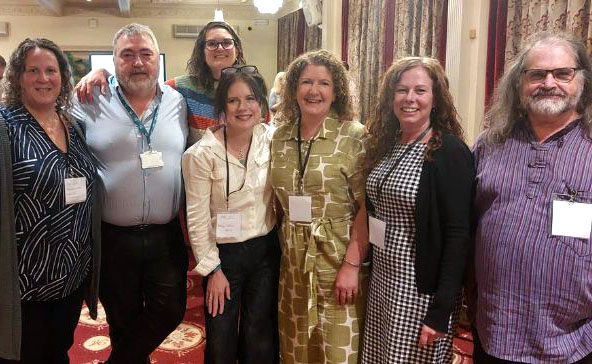 7 people in smart casual dress wearing lanyards standing side by side smiling at the camera.